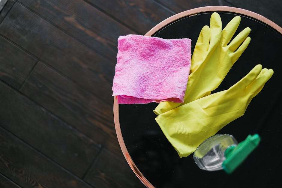 A round black surface table with a wooden edge, placed on a dark wooden floor. On the table, there is a pink microfiber cloth, a pair of yellow rubber cleaning gloves, and a small spray bottle with a