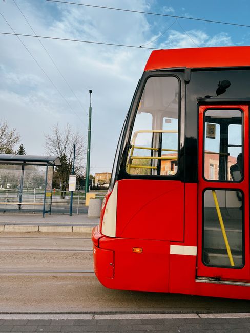A bright yellow tram is approaching a busy city tram stop outside an urban office building. The modern tram has large windows and is positioned on steel tracks embedded in the pavement. The station ar